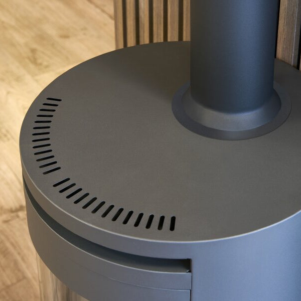 A close-up of a modern cylindrical Oko S5 bio ethanol metal heater / stove base with a dark gray top surface, vented slits, and a vertical chimney pipe, set against a wood slat wall and light wood floor.